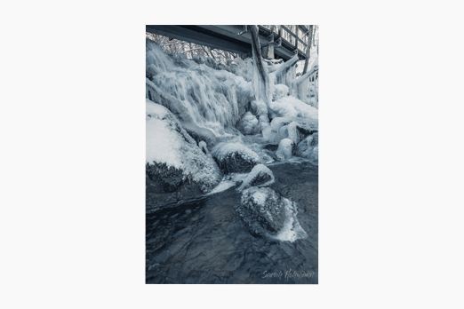 A frozen waterfall with ice bubbles and icicles. A bridge is going over the waterfall in Skåne in southern Sweden